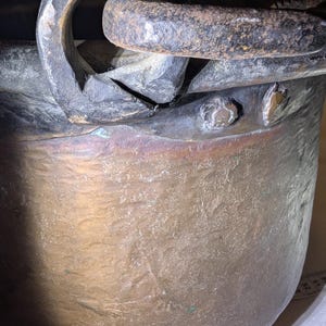 Può includere: Primo piano di un calderone antico in bronzo usurato con una grande maniglia curva. La superficie del calderone mostra segni di età e utilizzo, con una patina di colori. I rivetti fissano la maniglia alla pentola.
