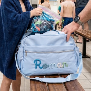 Könnte beinhalten: Eine hellblaue Sporttasche mit dem Namen "Roman" in blau und grüner Stickerei. Eine Person in einem blauen Handtuch hält eine Plastiktüte mit Schwimmausrüstung. Die Tasche hat einen Schultergurt und steht auf einer Holzbank.