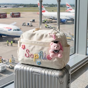 Peut inclure: Un sac de voyage crème brodé du nom "Sophie" en fil coloré, orné d'appliqués floraux et d'un charm en forme d'ours en peluche rose portant une toque de diplômé. Le sac est posé sur une valise à roulettes argentée, avec des avions en arrière-plan.