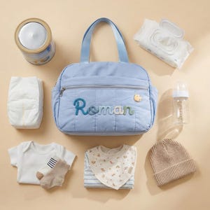 Könnte beinhalten: Hellblaue Wickeltasche mit dem Namen "Roman" aufgestickt. Das Bild zeigt Babyutensilien wie eine Windel, Feuchttücher, eine Flasche, Milchpulver, einen Body, Socken, ein Lätzchen und eine Strickmütze.