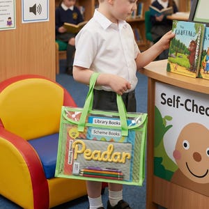 May include: A clear plastic book bag with bright green trim and handles, containing a gray pouch labeled Peadar in yellow script, a set of colored pencils, and two books labeled Library Books and Scheme Books, held by a child in a library.