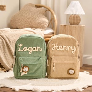 May include: Two corduroy backpacks, one sage green and one tan, are displayed on a white rug. The green backpack has the name "Logan" in white script, and a lion applique. The tan backpack has the name "Henry" in white script, and a lion applique.