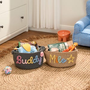 May include: Two woven pet baskets, one grey and one tan, sit on a jute rug. The grey basket has the name "Buddy" in rainbow letters, and the tan basket has "Milo" in rainbow letters. Each basket contains pet toys and treats.