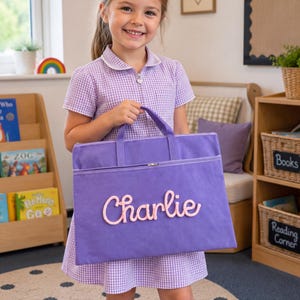 May include: A rectangular purple fabric book bag featuring two top handles and a horizontal zipper closure. The front of the bag displays the name Charlie in raised, light pink script lettering. The bag is constructed from a solid purple material.
