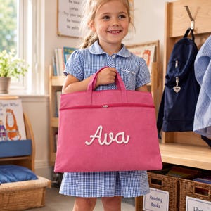 Peut inclure: Un sac à livres rectangulaire en toile rose doté de deux poignées supérieures et d'une fermeture éclair horizontale. Le devant du sac affiche le prénom Ava en lettres cursives blanches. Le sac est fabriqué dans un tissu rose uni.