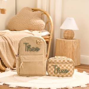 May include: A beige corduroy backpack and matching lunch bag, both personalized with the name "Theo" in green script. The backpack features a small cat face patch. The lunch bag has a checkered pattern.
