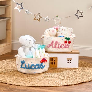 May include: Two cream-coloured rope baskets, labelled "Lucas" in blue and "Alice" in pink, filled with baby essentials. The Lucas basket contains a teddy bear, nappies, and a toy car. The Alice basket holds a stuffed bunny and floral-patterned items.