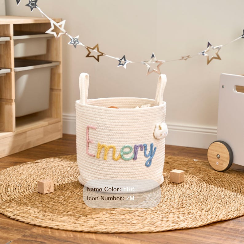 May include: A cream-coloured, woven rope storage basket with the name "Emery" embroidered in colourful letters. The basket has two handles and a small bunny applique. The basket sits on a round, woven mat.