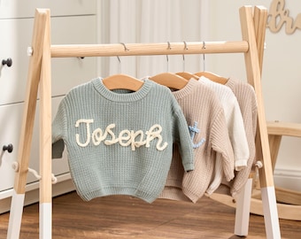 May include: A wooden clothing rack displays several knit sweaters on hangers. The sweaters are in shades of blue, beige, and cream. One sweater has the name "Joseph" in a cream-colored script. The rack has white-painted legs.