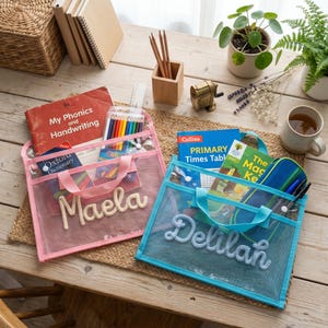 May include: Two rectangular mesh zipper pouches with handles, one pink and one blue. The pink pouch features the name Maela in white yarn lettering, while the blue pouch features the name Delilah in light blue yarn lettering. Both are transparent.