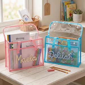 May include: Two rectangular clear plastic storage bags with handles and snap closures. The left bag has pink trim and the name Maela in script. The right bag has blue trim and the name Delilah in script. Both bags contain notebooks and pens.
