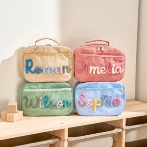 Puede incluir: Cuatro bolsas de almuerzo de pana en beige, rosa, verde y azul, cada una con un nombre diferente bordado en la parte delantera. Los nombres son Roman, Amelia, Wilson y Sophie. Cada bolsa tiene un asa superior y cierre de cremallera.
