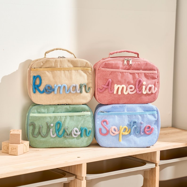Puede incluir: Cuatro bolsas de almuerzo de pana en beige, rosa, verde y azul, cada una con un nombre diferente bordado en la parte delantera. Los nombres son Roman, Amelia, Wilson y Sophie. Cada bolsa tiene un asa superior y cierre de cremallera.