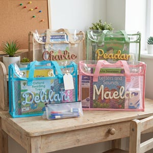 May include: Four rectangular clear mesh tote bags with coloured fabric handles and trim. Each bag features a personalised name in raised yarn lettering on the front. The bags contain various books, stationery, and a small clear pencil case with pens.