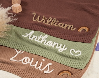 Pode incluir: Uma pilha de três mantas de malha personalizadas em tons de castanho, verde e bege. Cada manta apresenta um nome bordado em escrita branca: William, Anthony e Louis. Os elementos decorativos incluem um arco-íris e um coração.