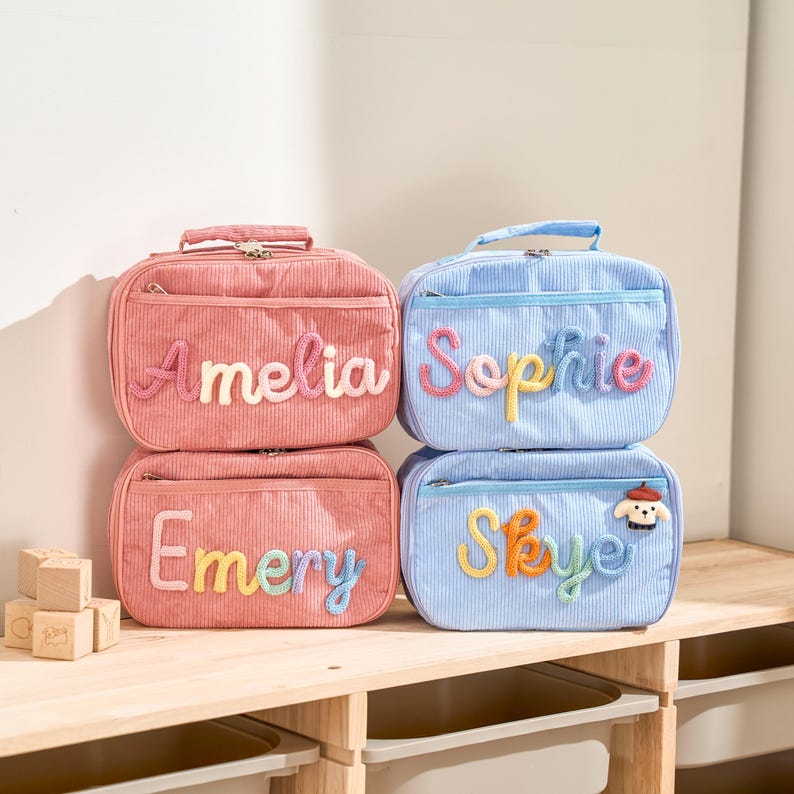 Puede incluir: Cuatro loncheras personalizadas en pana rosa y azul. Las bolsas rosas est&aacute;n etiquetadas con "Amelia" y "Emery" con letras cosidas de colores. Las bolsas azules est&aacute;n etiquetadas con "Sophie" y "Skye" con letras cosidas y un parche de perro.
