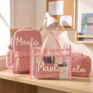 May include: A pink corduroy backpack, a clear plastic tote bag with pink trim, and a pink corduroy pencil case. The backpack and pencil case feature the name Maela in white script. The clear tote contains a notebook, pens, and a calculator.