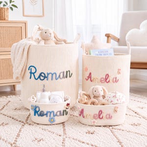 Peut inclure: Deux grands paniers de rangement en velours côtelé crème et deux petits paniers en corde tressée, personnalisés avec les noms brodés Roman ou Amelia. Les paniers contiennent des peluches, un livre intitulé Guess How Much et divers articles.