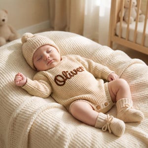 Peut inclure: Une barboteuse à manches longues en tricot couleur crème avec le prénom Oliver brodé en lettres cursives marron sur la poitrine. La tenue comprend un bonnet côtelé assorti avec un pompon et des chaussons à nouer, avec deux petits boutons en bois.