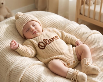 May include: A cream-coloured knitted long-sleeve romper with the name Oliver embroidered in brown script across the chest. The outfit includes a matching ribbed beanie with a pom-pom and booties with tie closures, featuring two small wooden buttons.