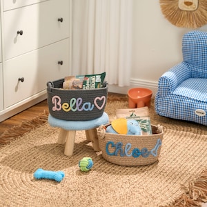 May include: Two pet baskets, one grey with the name "Bella" in rainbow colours and a heart, the other tan with "Chloe" in blue. The baskets sit on a woven rug with pet toys and a blue gingham chair in the background.
