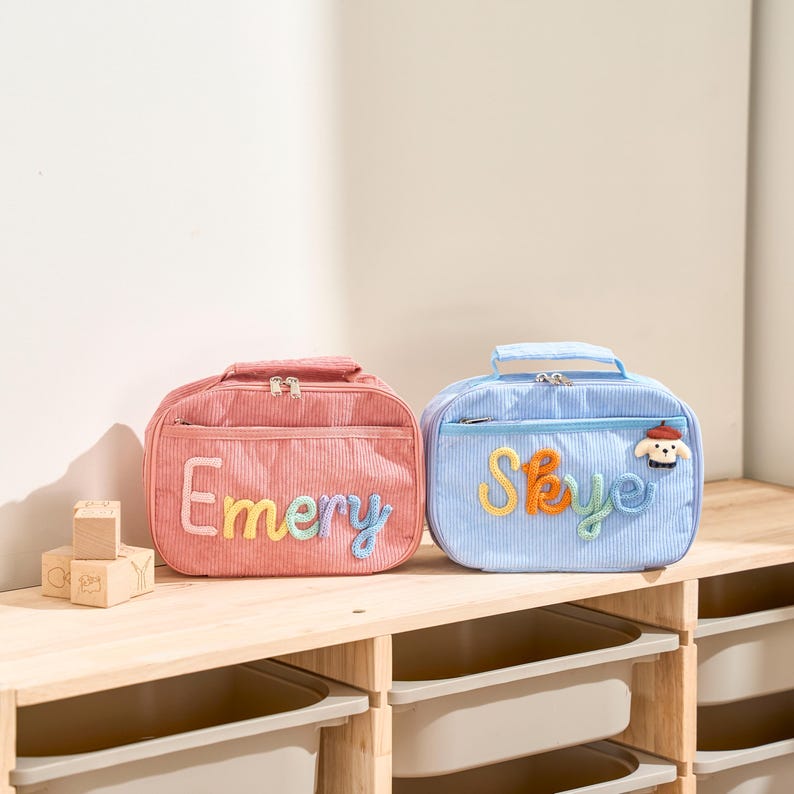 Puede incluir: Dos loncheras de pana, una rosa y otra azul, sobre una estanter&iacute;a de madera. La bolsa rosa dice "Emery" en letras cosidas de colores. La bolsa azul dice "Skye" con un parche de perro. Ambas tienen asas superiores y cremalleras.