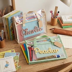 May include: Two rectangular transparent mesh zipper pouches with beige and light green fabric trim. The beige pouch features the name Charlie in pink yarn, and the light green pouch features the name Peadar in yellow yarn. Both include blank brown tags.