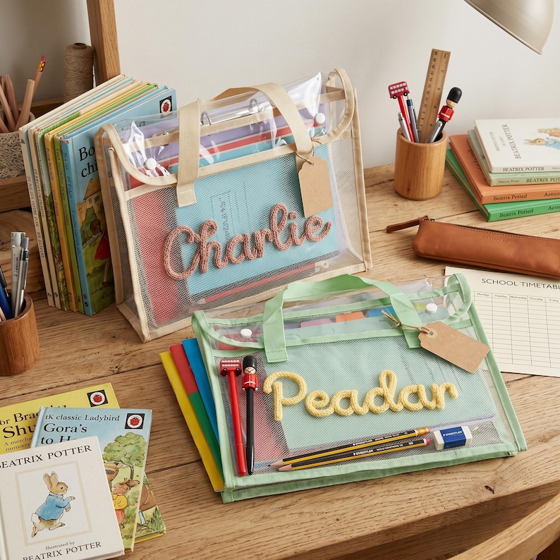 May include: Two rectangular transparent mesh zipper pouches with beige and light green fabric trim. The beige pouch features the name Charlie in pink yarn, and the light green pouch features the name Peadar in yellow yarn. Both include blank brown tags.