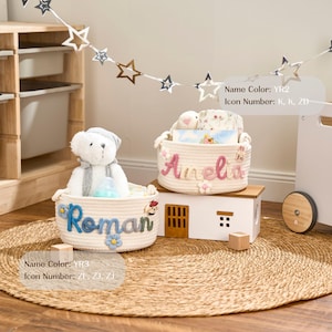 May include: Two cream rope baskets, one with "Roman" in blue and the other with "Amelia" in pink, sit on a woven mat. The "Roman" basket contains a teddy bear. The "Amelia" basket has a bunny. Both are filled with toys and are decorative.