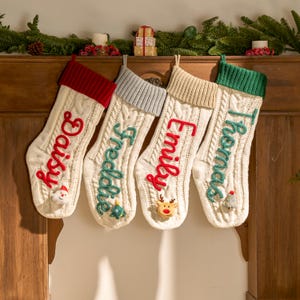 May include: Four cable-knit Christmas stockings with personalized names: "Daisy" in red, "Freddie" in teal, "Emily" in red, and "Thomas" in teal. Each stocking has a different colored cuff and a small decorative element at the toe.