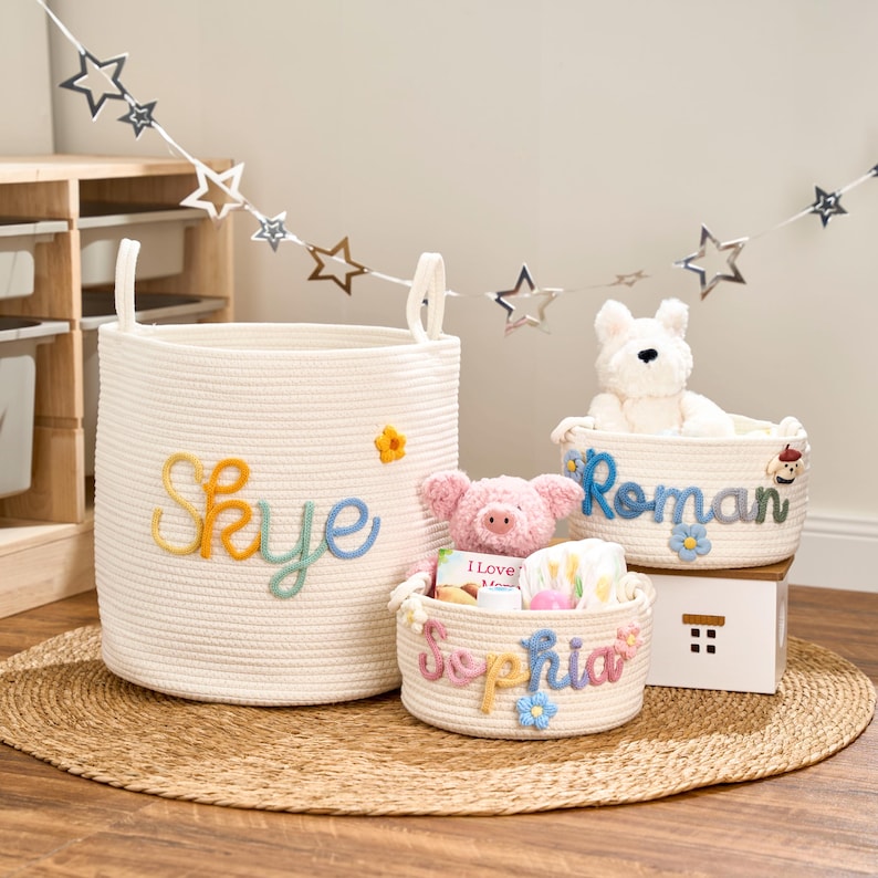 Op de afbeelding: Drie witte touwmanden met geborduurde namen en bloemaccenten. De grootste mand heeft de naam "Skye" in regenboogkleuren. De andere twee manden hebben de namen "Roman" en "Sophia". Een knuffelvarken en een hond zitten in de manden.