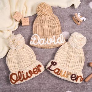 May include: Three knitted beanies with pom-poms. One is tan with "David" in white, and two are cream-coloured with "Oliver" in brown and "Lauren" in brown, orange, and yellow. The hats are on a grey surface with wooden toys.