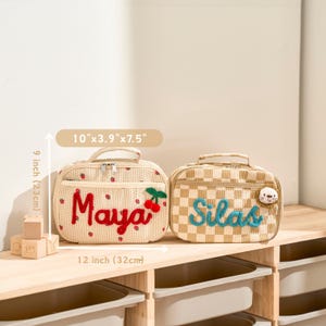 Puede incluir: Dos bolsas de almuerzo personalizadas. La bolsa de la izquierda es beige con detalles de fresas rojas y el nombre "Maya" en rojo. La bolsa de la derecha es a cuadros beige y blanco con el nombre "Silas" en azul. Cada bolsa mide 25cm x 10cm x 19cm.
