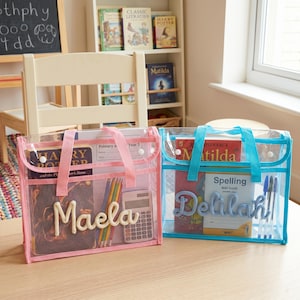 May include: Two rectangular clear plastic document bags with handles and snap closures. The left bag is trimmed in pink with the name Maela in cream yarn. The right bag is trimmed in blue with the name Delilah in blue yarn. Both contain books and supplies.