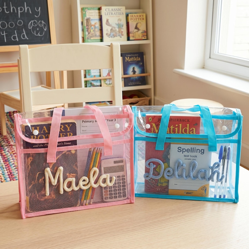 May include: Two rectangular clear plastic document bags with handles and snap closures. The left bag is trimmed in pink with the name Maela in cream yarn. The right bag is trimmed in blue with the name Delilah in blue yarn. Both contain books and supplies.