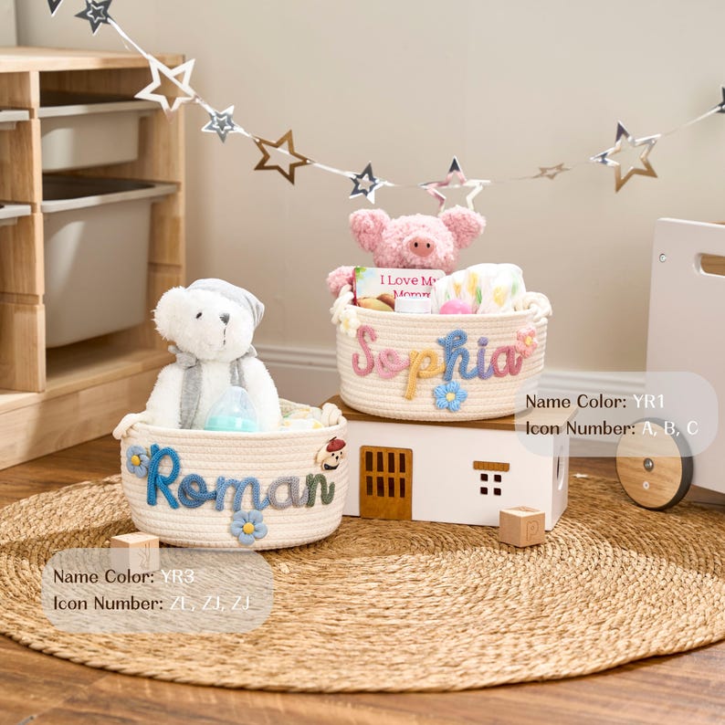 Op de afbeelding: Twee cr&egrave;mekleurige touwmanden met gepersonaliseerde namen en bloemaccenten. E&eacute;n mand heeft de naam "Roman" in blauw, de andere "Sophia" in roze. Elke mand bevat een knuffel en andere items. De manden staan op een ronde geweven mat.