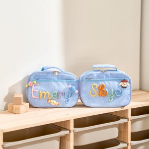 May include: Two light blue lunch bags with corduroy texture, each with a handle and zipper. One bag has the name "Emery" in colorful letters and candy accents. The other bag has the name "Skye" and a dog patch.