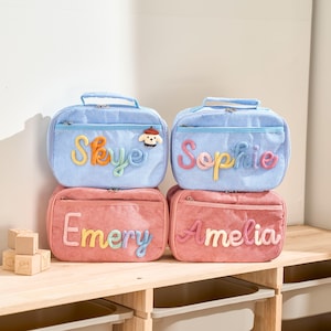 Peut inclure: Quatre sacs à lunch personnalisés en velours côtelé bleu clair et rose corail. Les deux sacs supérieurs sont bleu clair avec les noms "Skye" et "Sophie" brodés en fil coloré. Les deux sacs inférieurs sont rose corail avec les noms "Emery" et "Amelia".