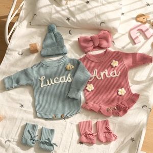 Könnte beinhalten: Ein Baby-Kleidungsset mit einem hellblauen Strick-Strampler mit "Lucas" in weißer Schrift, passendem Hut und Booties. Ein rosa Strampler mit "Aria" in weißer Schrift, ein Stirnband und Booties sind ebenfalls abgebildet. Die Outfits sind mit kleinen Blumen verziert.