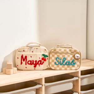 Könnte beinhalten: Zwei personalisierte Lunchpakete. Eines ist beige mit roten Erdbeerakzenten und dem Namen "Maya" in roter Schrift, mit einem Kirschakzent. Das andere ist tan und weiß kariert mit dem Namen "Silas" in blauer Schrift und einem kleinen Tieraufnäher.