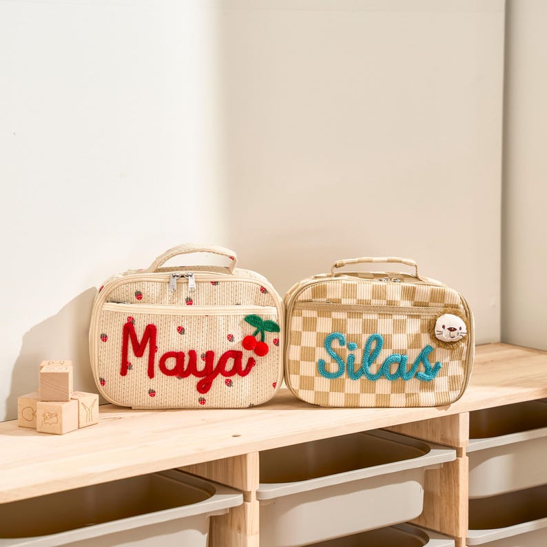 Puede incluir: Dos loncheras personalizadas. Una es beige con detalles de fresas rojas y el nombre "Maya" en escritura roja, con un acento de cereza. La otra es a cuadros beige y blancos con el nombre "Silas" en escritura azul y un peque&ntilde;o parche de animal.
