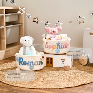 May include: Two cream rope baskets, each with a name: "Roman" and "Sophia", adorned with flowers. One basket holds a teddy bear and baby bottle, the other a pig and baby items. They sit on a woven mat, with a wooden toy house in the background.