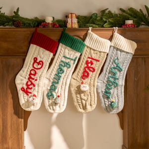 May include: Four cable-knit Christmas stockings with personalized names: "Daisy" in red, "Thomas" in green, "Isla" in red, and "Arthur" in teal. Each stocking has a different colored cuff and a small decorative element.
