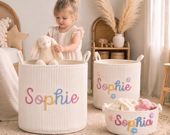 May include: Three cylindrical off-white storage baskets made of thick corded fabric. Each basket features the name Sophie embroidered in colourful, raised script lettering, accented with small, pastel-coloured fabric flowers attached to the sides.