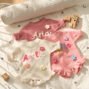 May include: Three baby sweaters in shades of pink and white, decorated with floral details and personalised names. The sweaters have short sleeves, a round neck, and a frilled hem. The names "Aria" and "Amelia" are embroidered on the sweaters.