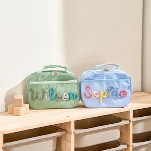 Puede incluir: Dos loncheras de pana, una verde y otra azul, est&aacute;n sobre una estanter&iacute;a de madera. La bolsa verde tiene el nombre "Wilson" en hilo colorido, y la bolsa azul tiene "Sophie". Ambas tienen un asa y una cremallera.
