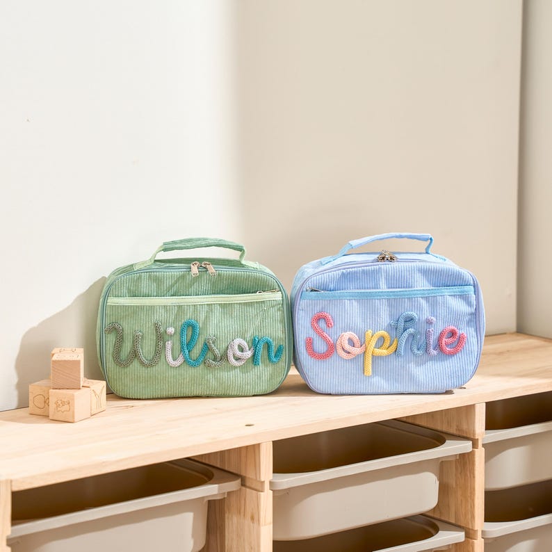 Puede incluir: Dos loncheras de pana, una verde y otra azul, est&aacute;n sobre una estanter&iacute;a de madera. La bolsa verde tiene el nombre "Wilson" en hilo colorido, y la bolsa azul tiene "Sophie". Ambas tienen un asa y una cremallera.