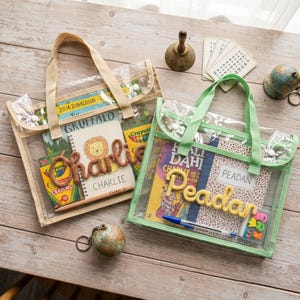 May include: Two rectangular clear mesh tote bags with fabric handles. The left bag contains a book, a box of crayons, and a notebook with the name Charlie in brown yarn. The right bag contains a book, a notebook with the name Peadan in yellow yarn, a blue pen, and a small box of erasers.