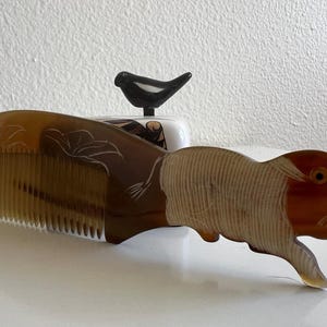 May include: A decorative comb with a fish-shaped handle. The comb is made of a light brown material with carved floral details. The fish handle is a darker brown with a carved design. A small black bird figurine sits behind the comb.