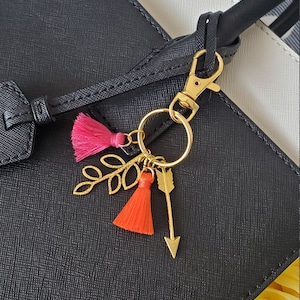 May include: A gold key chain with a pink and orange tassel charm, a gold arrow charm, and a gold leaf charm. The keychain is attached to a black leather handbag.
